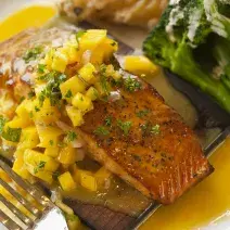Fotografia em tons de amarelo em uma bancada de madeira de cor marrom. Ao centro, um prato branco contendo o peixe com alguns legumes ao lado.