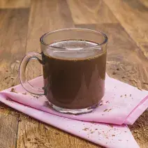 Fotografia de uma bancada de madeira com um paninho rosa e uma xícara transparente com o chocolate quente dentro.