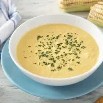Fotografia em tons de amarelo e azul em uma bancada de madeira clara, um prato azul e branco ao centro com sopa de milho. Ao lado uma espiga de milho e um paninho azul quadriculado.