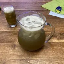 Fotografia em tons de azul e verde em uma bancada de madeira com paninhos verde e azul ao fundo. Ao centro, uma jarra de vidro grande com o chá de limão com abacaxi e hortelã e ao lado um copo com a bebida.