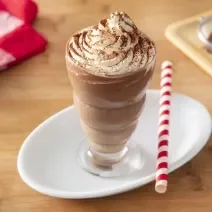 Foto da receita de Milkshake de Cappuccino. Observa-se um copo alto com a bebida decorado com chantilly e polvilhado de chocolate em pó. Ao lado direito, um canudo vermelho e branco para decorar.