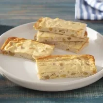 Foto em tons de amarelo da receita de barrinha proteica de cheesecake de maçã servida em diversas porções sobre uma panela branca de cerâmica em cima de uma mesa de madeira azul com paus de canela e maçã