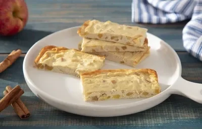 Foto em tons de amarelo da receita de barrinha proteica de cheesecake de maçã servida em diversas porções sobre uma panela branca de cerâmica em cima de uma mesa de madeira azul com paus de canela e maçã