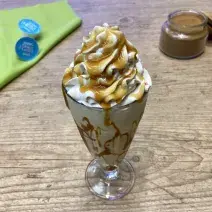 Fotografia em tons de azul e verde em uma bancada de madeira e um copo de vidro ao centro com a bebida de Milkshake e ao fundo um potinho com canela em pó.