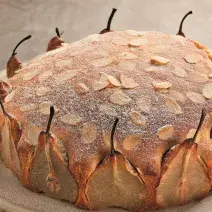 Fotografia em tons de cinza e marrom de uma bancada cinza vista de cima, um recipiente redondo contém um bolo com peras cortas em volta do bolo para decorar