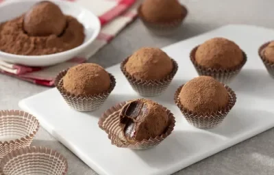 Fotografia em tons de branco, cinza e marrom de uma bancada cinza com um prato retangular branco com trufas de chocolate zero em forminhas marrom. Ao fundo um paninho xadrez branco e vermelho, um recipiente branco redondo com chocolate em pó e uma trufa.