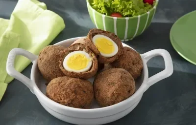 Foto da receita de Bolovo. Observa-se um recipiente com 6 bolovos, sendo que um está cortado no meio. Atrás, um recipiente com salada decora. Foto da receita de Bolovo. Observa-se um recipiente com 6 bolovos, sendo que um está cortado no meio. Atrás, um recipiente com salada decora.