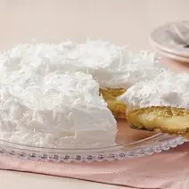 Fotografia de um bolo coberto com marshmallow branco e raspada de coco em cima. Dentro do bolo creme e massa brancos. O prato está em cima de um guardanapo de papel rosa claro.