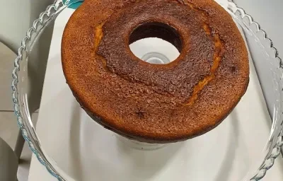 Foto da receita de bolo de cuscuz servida em uma porção grande sobre uma bailarina de vidro em cima de uma mesa branca