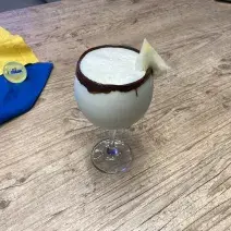 Fotografia em tons de branco, amarelo e azul em uma bancada de madeira com um pano azul, amarelo e uma taça de vidro com a bebida de Galak dentro e uma fatia de abacaxi decorando.