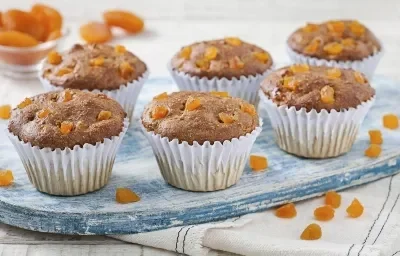 Fotografia em tons de azul em uma bancada de madeira clara, um paninho branco com listras azuis escuras, uma tábua de madeira azul e vários muffins integrais com damasco em forminhas brancas.