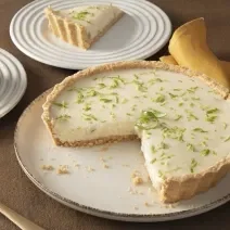 Foto da receita de torta de limão com chandelle servida em uma porção grande sobre uma base de porcelana branca com um pedaço cortado em um pratinho menor ao lado. À frente há uma espátula e ao fundo um pano amarelo