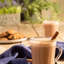 Fotografia em tons de marrom em uma bancada de madeira com um pano azul e duas xícaras de vidro com o chocaccino e um pau de canela para enfeitar. Ao fundo, um prato com cookies