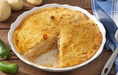 Foto em tons de amarelo da receita de batata apimentada com queijo servida em uma porção grande em uma forma branca sobre uma mesa de madeira com um pano azul quadriculado Foto em tons de amarelo da receita de batata apimentada com queijo servida em uma porção grande em uma forma branca sobre uma mesa de madeira com um pano azul quadriculado