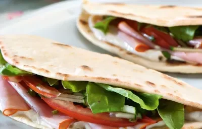 Fotografia em tons de rosa em uma mesa de madeira com um pano rosa florido, uma tábua de madeira branca ao centro com a piadina, que é uma massa estilo crepe francês, recheada de tomate, rúcula e peito de peru. Fotografia em tons de rosa em uma mesa de madeira com um pano rosa florido, uma tábua de madeira branca ao centro com a piadina, que é uma massa estilo crepe francês, recheada de tomate, rúcula e peito de peru.