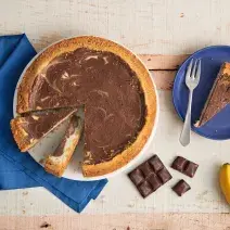Fotografia em tons de azul em uma bancada de madeira, um pano azul, um prato grande raso com a cheesecake de banana com chocolate em cima dele. Ao lado, um prato pequeno azul raso com uma fatia da cheesecake, um cacho de banana e quadradinhos de chocolate