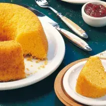 Fotografia em tons de amarelo, branco e azul escuro de uma bancada azul escura vista de cima. Um prato redondo e branco com o bolo faltando uma fatia e ao lado um prato redondo branco com uma fatia de bolo.