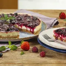 Fotografia em tons de vermelho em uma bancada de madeira com um prato raso grande com a cheesecake de iogurte com frutas vermelhas e um prato raso pequeno com uma fatia da cheesecake. Morangos e uvas espalhadas pela bancada.
