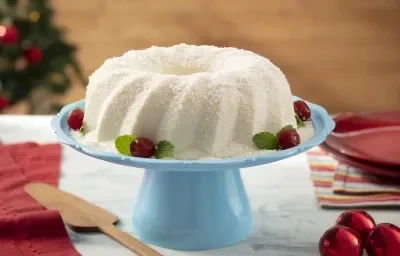 Foto da receita de Sobremesa Gelada de Coco. Observa-se uma boleira azul com a sobremesa em cima, polvilhada de coco e enfeitada com cerejas na base. Decorações de Natal enfeitam a foto, como a árvore no fundo desfocada e bolas natalinas.