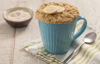 Fotografia em tons de branco e azul de uma bancada de madeira branca com um paninho verde, branco e azul, sobre ele uma caneca azul com bolo de caneca e uma colher. Ao fundo um recipiente redondo com aveia Naturnes.