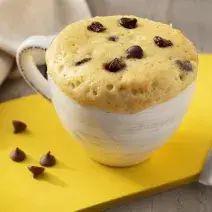 Fotografia em tons de marrom. branco e amarelo de uma bancada de madeira com uma tábua amarela, sobre ela uma xícara branca com bolo de caneca e uma colher. Ao fundo um paninho branco.