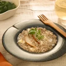 Foto da receita de risoto de pera com brie servida em um prato fundo azul com um garfo de madeira na borda e um copo de vidro com vinho branco ao fundo