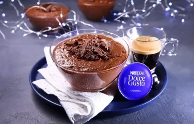 Fotografia em tons de azul em uma bancada de cimento queimado, um prato redondo azul com uma taça de vidro com a mousse de chocolate dentro e um paninho branco ao lado. Ao lado, uma xícara de vidro com café e atrás decoração com luzes natalina.