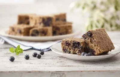 Fotografia em tons de azul, marrom e branco, tendo à frente prato branco com pedaços de bolo com blueberry e blueberries espalhadas na bancada, ao fundo outro prato com pedaços de bolo sobre guardanapo branco de borda azul, tudo sobre bancada branca.