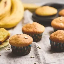 Fotografia de quatro muffins de banana e aveia dentro de uma forminha de papel colorida sobre uma toalha de mesa em tons de bege. Ao lado, um cacho de bananas, duas fatias do bolo, e ao fundo, dois muffins em uma forma preta de bolinhos.