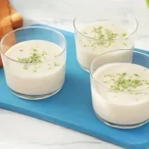 Foto da receita de mousse de limão zero lactose servida em três porções em taças sobre uma tábua azul com um pano laranja ao lado além de duas colheres prateadas