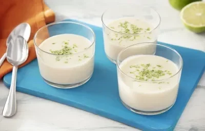 Foto da receita de mousse de limão zero lactose servida em três porções em taças sobre uma tábua azul com um pano laranja ao lado além de duas colheres prateadas Foto da receita de mousse de limão zero lactose servida em três porções em taças sobre uma tábua azul com um pano laranja ao lado além de duas colheres prateadas