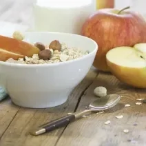 Fotografia de um recipiente fundo com maçã em pedaços, cereais e avelãs por cima. Ao lado, uma colher de sobremesa, e mais ao fundo, uma maçã inteira e outra cortada pela metade, sobre uma mesa de madeira.