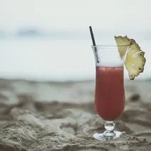 Fotografia de uma taça com um coquetel de água com gás e frutas, e na borda, metade de uma fatia de abacaxi. Dentro da bebida tem um canudo preto, na qual está apoiada sobre a areia.