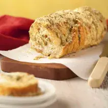 Fotografia em tons amarelo e vermelho em uma bancada de madeira clara, uma tábua de madeira com um papel toalha e o pão de cebola em cima dele. Ao lado, uma faca de pão e uma fatia do pão em cima de um pratinho branco pequeno.
