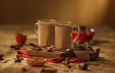 Fotografia em tons de marrom em uma bancada de madeira de cor marrom. Ao centro, um pano vermelho contendo 2 xícaras com o chocolate quente e ao redor há alguns pedações de chocolates espalhados.