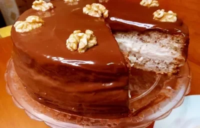 Foto em tons de marrom da receita de bolo de nozes festivo servida em uma porção grande com uma fatia retirada, mostrando o interior do bolo com o recheio. Está sobre uma bailarina (base utilizada na cozinha) em cima de uma mesa de madeira