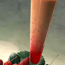 Fotografia em tons de vermelho em uma bancada de madeira, várias frutas vermelhas espalhadas e um copo de vidro super grande com a bebida feita com pitanga e Leite Moça dentro dele.