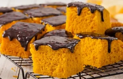 Fotografia em tons de amarelo e preto de uma bancada de madeira com um potinho branco com calda de chocolate, ao lado uma grade preta com fatias quadradas de bolo de cenoura com calda de chocolate em cima, ao fundo, um pano amarelo.