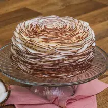 Foto aproximada da receita de Bolo de Baba de Moça com Nozes, decorado com fitas de coco, sobre um prato de vidro em cima de um pano cor de rosa, numa bancada de madeira com um pedaço de coco.