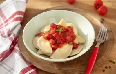 Foto da receita de Pierogi (massa polonesa). Observa-se um bowl verde claro com a massa dentro, servida de molho de tomate, sobre uma tábua de madeira escura com um garfo ao lado direito.