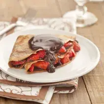 Fotografia em tons de marrom em uma bancada de madeira com um pano branco com listras marrom, um prato branco raso com o crepe recheado com morangos e chocolate.