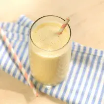 Foto de uma bancada de madeira clara. Sobre ela há uma pano listrado azul e branco e, apoiado nele, um copo com a bebida amarela, um canudo de papel laranja e branco dentro do copo e outro sobre o pano.