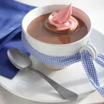 Fotografia em tons de azul em uma bancada de madeira branca com um pano azul ao lado, um prato branco raso pequeno, um xícara branca e o chocolate quente com marshmallow dentro.