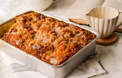 Foto da receita de Cuca de Banana com Leite MOÇA. Observa-se um refratário retangular com a cuca dentro, crocante e dourada com calda de Leite MOÇA. Ao lado direito, um recipiente de servir calda de cerâmica.