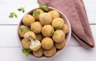 Fotografia em tons de branco e rosa de uma bancada branca, sobre ela um recipiente rosa com bolinhos de arroz e queijo. Ao lado um paninho rosa.