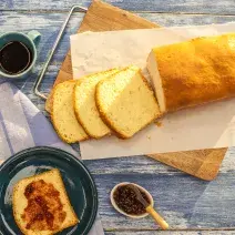 Fotografia em tons de azul e branco de uma bancada azul com uma tábua, sobre ela um paninho branco com o pão. Ao lado um paninho branco e azul, sobre ele um prato redondo azul com uma fatia de pão e geleia e uma xícara de café.