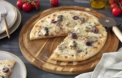 Foto da receita de pizza de sardinha servida em uma tábua de madeira circular com um dos pedaços separados em um prato de porcelana branco à frente. Para a decoração, há tomatinhos cereja, um paninho cinza, um vidro de azeite e um cortador de pizza