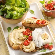 fotografia em tons de bege, branco e verde de uma bancada bege vista de cima. Contém um prato retangular branco com  fatias de pão, homus e tomates.