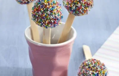 Fotografia em tons de lilás e azul, com copo lilás contendo três pirulitos de brigadeiro com confeitos coloridos, com mais um pirulito ao lado sobre guardanapo colorido, tudo sobre bancada em tons de azul. Fotografia em tons de lilás e azul, com copo lilás contendo três pirulitos de brigadeiro com confeitos coloridos, com mais um pirulito ao lado sobre guardanapo colorido, tudo sobre bancada em tons de azul.