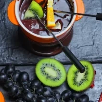 Fotografia de um Fondue de chocolate meio amargo dentro de um recipiente de vidro na cor laranja, com frutas sobre o creme. Ao lado, algumas frutas sobre uma mesa preta, como uvas roxas e um kiwi cortado na metade.